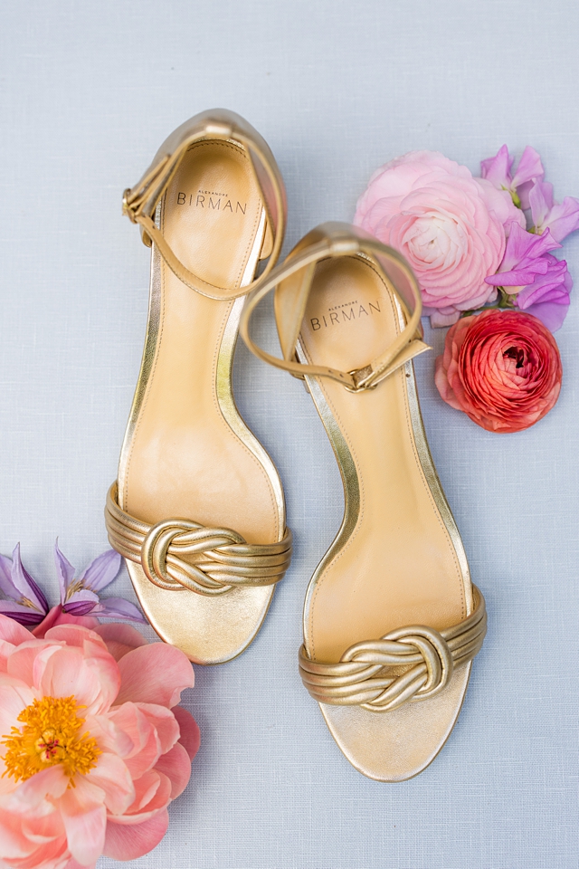Gold wedding shoes by Birman sitting on blue background with colorful florals around shoes